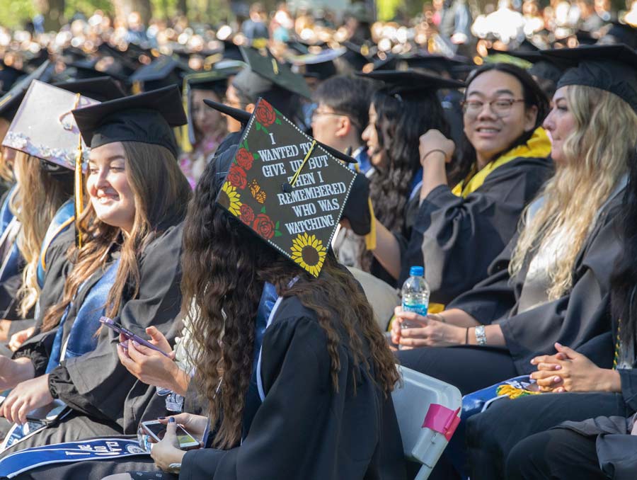 A group of students sitting at graduation. One student's graduation cap is decorated and reads, "I wanted to give up but then I remembered who was watching."
