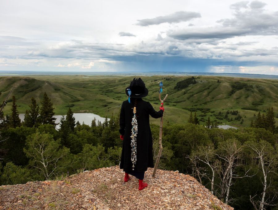 The back of a person clad in black on a hilltop holding a cane looking out at a cloudy horizon.