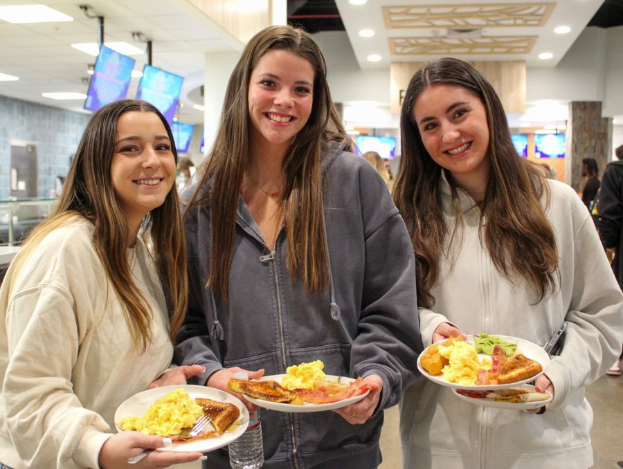 Three students holding plates of food smiling at camera at Pack Place, dining hall.
