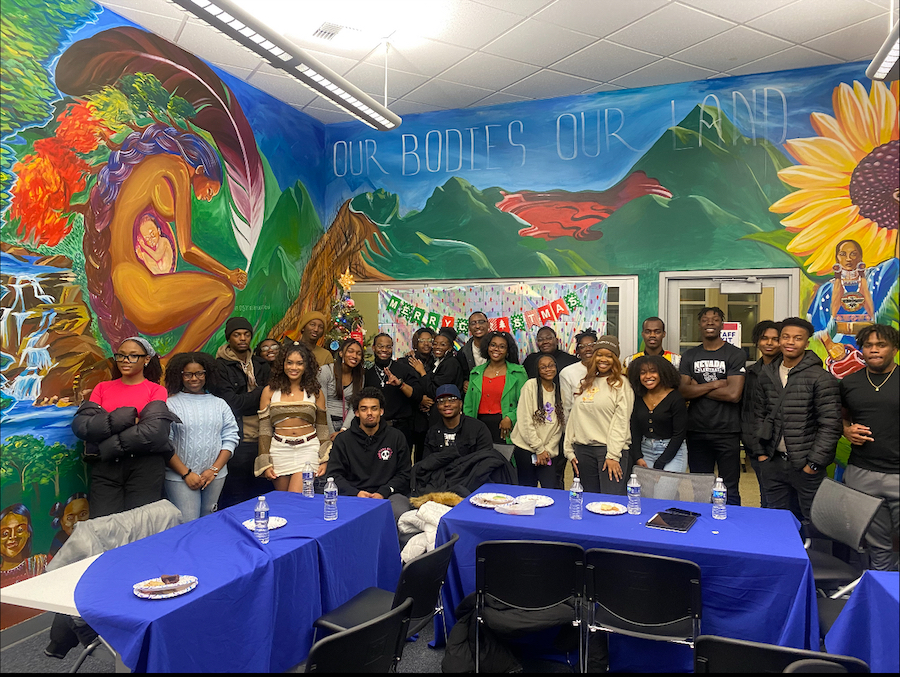 A group of 24 students stand in front of two tables in the Multicultural Center on campus. The wall behind them is a mural with flowers and green hills and the words "Our Bodies Our Land" is visible in the mural.