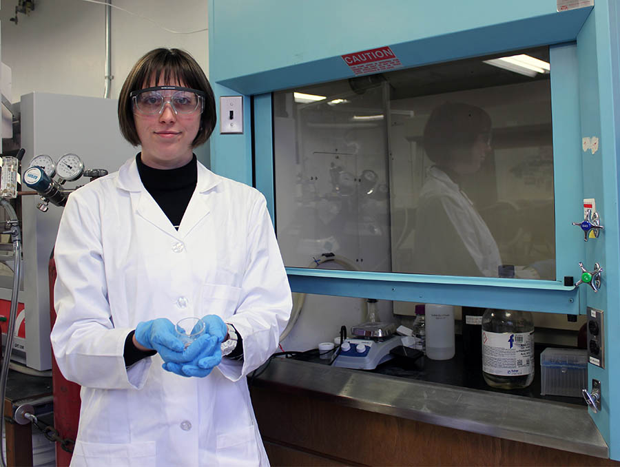 Harmony Werth, wearing a lab coat, gloves and eye protection, stands in Krista Carlson's lab holding a small clear container in both hands. 