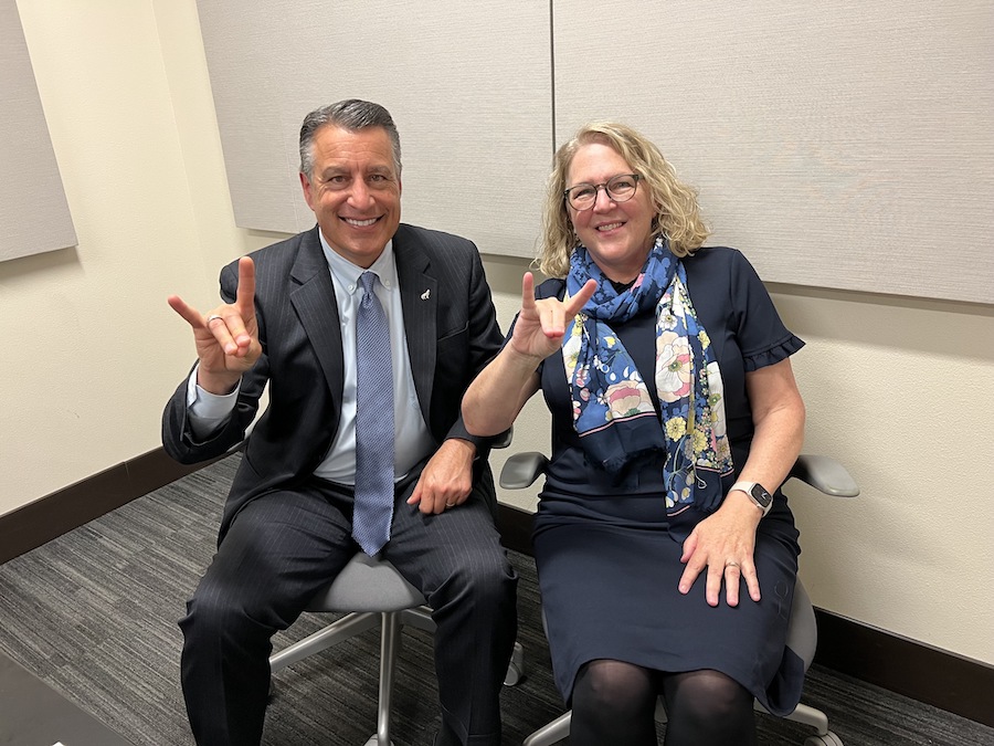 President Brian Sandoval sits next to Catherine Cardwell in a podcasting studio. Both people hold their hands in the wolf sign.