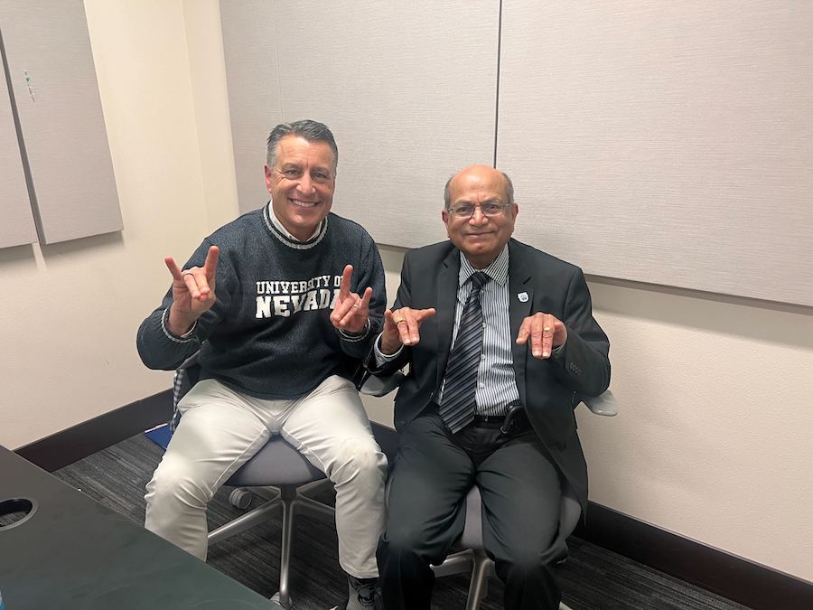 President Brian Sandoval sits next to Sasi Pillay in a podcasting studio. Both people hold their hands in the wolf sign.