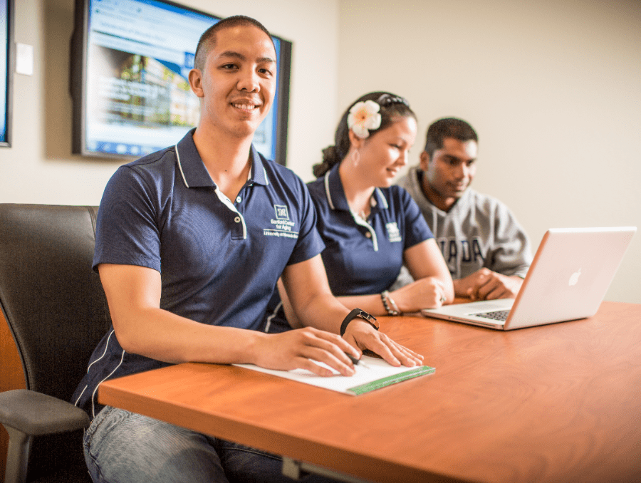 Three Gerontology Students sitting at a desk, wearing branded Sanford polo shirts. Two look at a laptop.