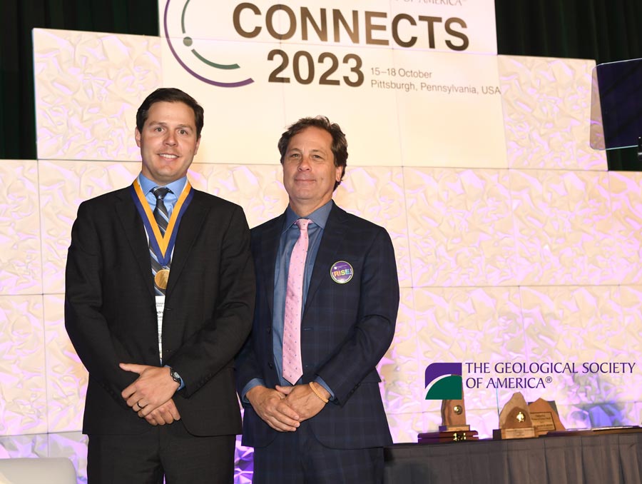 Andrew Zuza smiles, wearing a medal and standing next to another man who is smiling on a stage.
