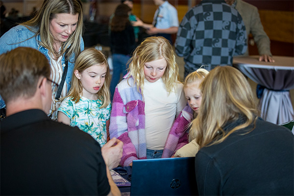 Three young girls and and an adult woman look at a computer screen on a display table.