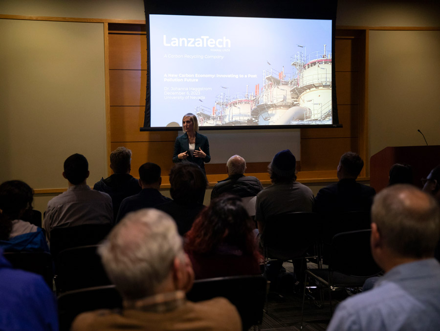 A woman stands in front of an audience with a presentation being projected onto a screen behind her.