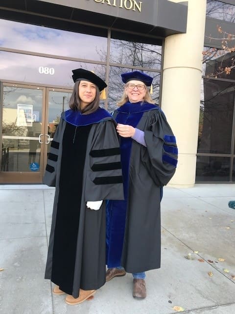 Two women stand and smile while wearing hooding graduation regalia on campus.