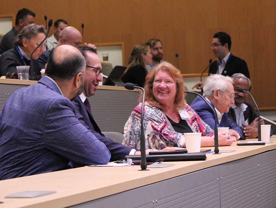 Five people seated and talking at a long table in an amphitheater style conference room, with seven people behind them talking amongst themselves. 