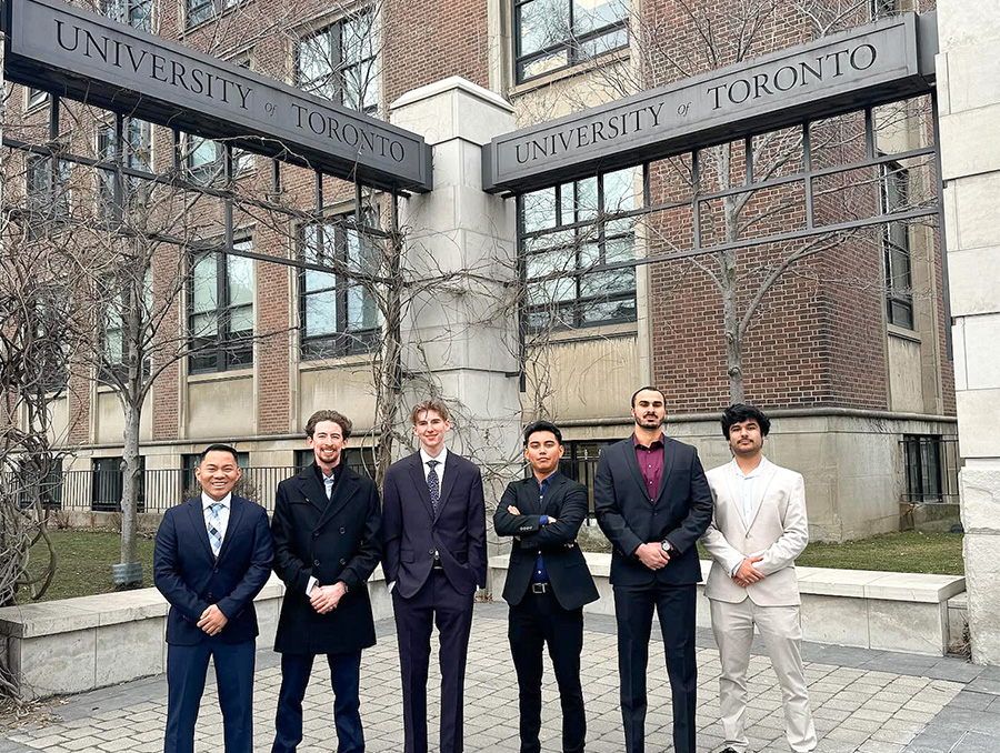 Six teammates pose in front of University of Toronto sign wearing business suits.