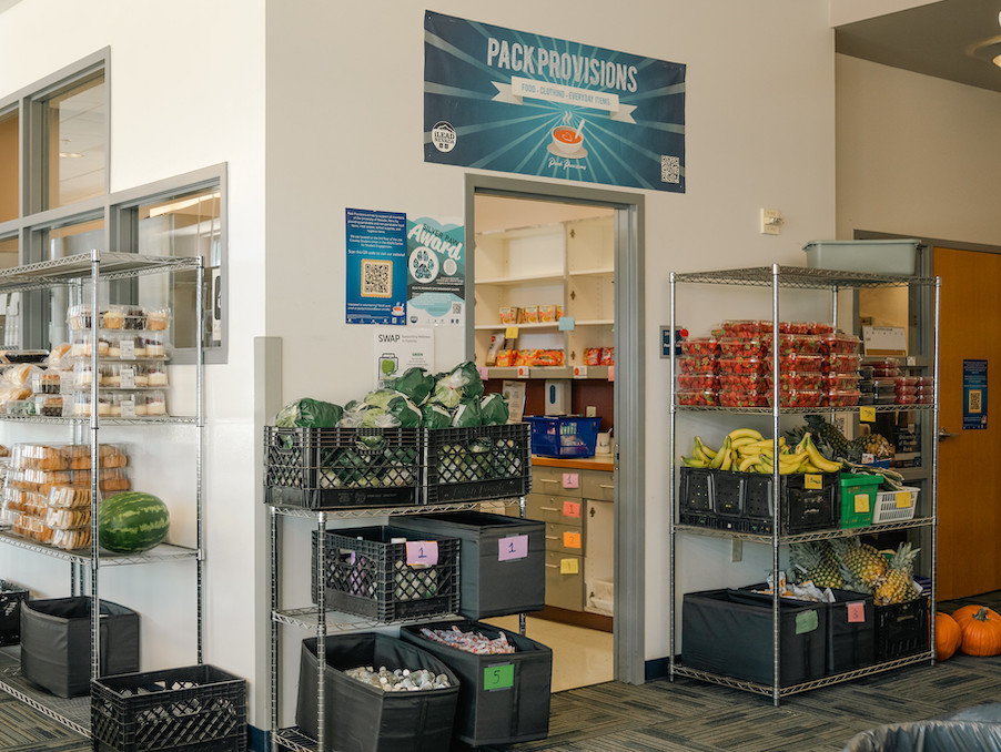 The Pack Provisions food pantry entrance with a Pack Provisions sign above the door and shelves of food in the hallways including fresh fruits and vegetables.