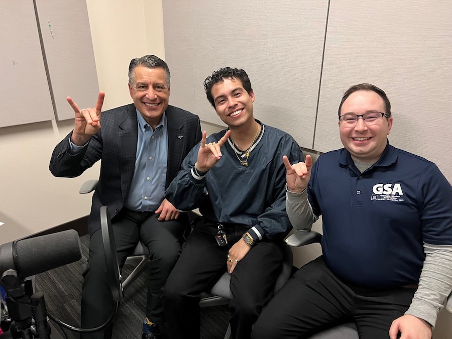 Brian Sandoval sitting next to Boris Carpio Guerra and Matthew Hawn in the podcasting studio holding up wolf pack hand signs.
