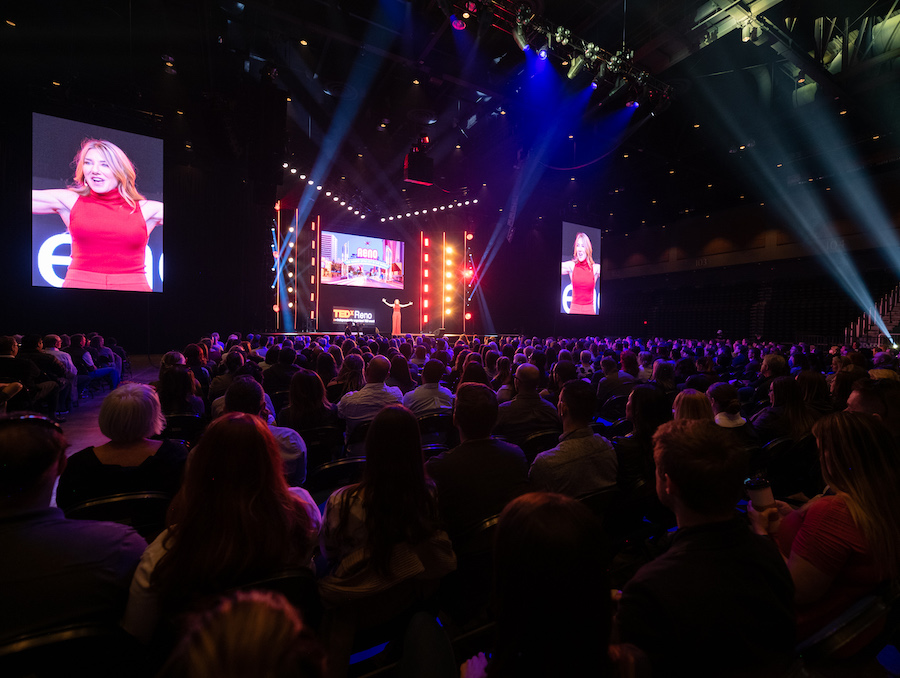 A large audience sits in front of the TEDxReno stage while a woman stands giving a talk and appears on two large screens on either side of her.