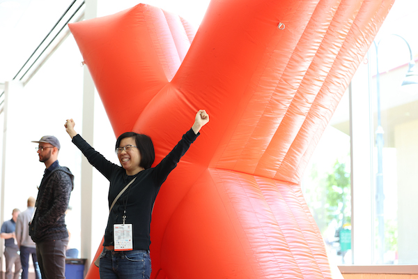 A woman wears and name badge and poses with her hands in the air in front of an inflatable X.