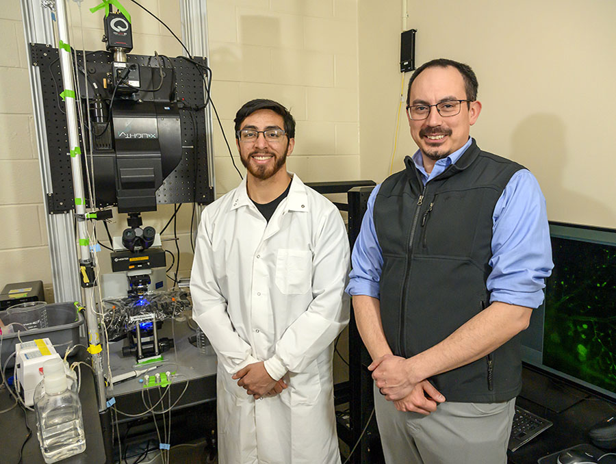 Cristian Franco and Albert Gonzales pose for a photo in front of a lab microscope.