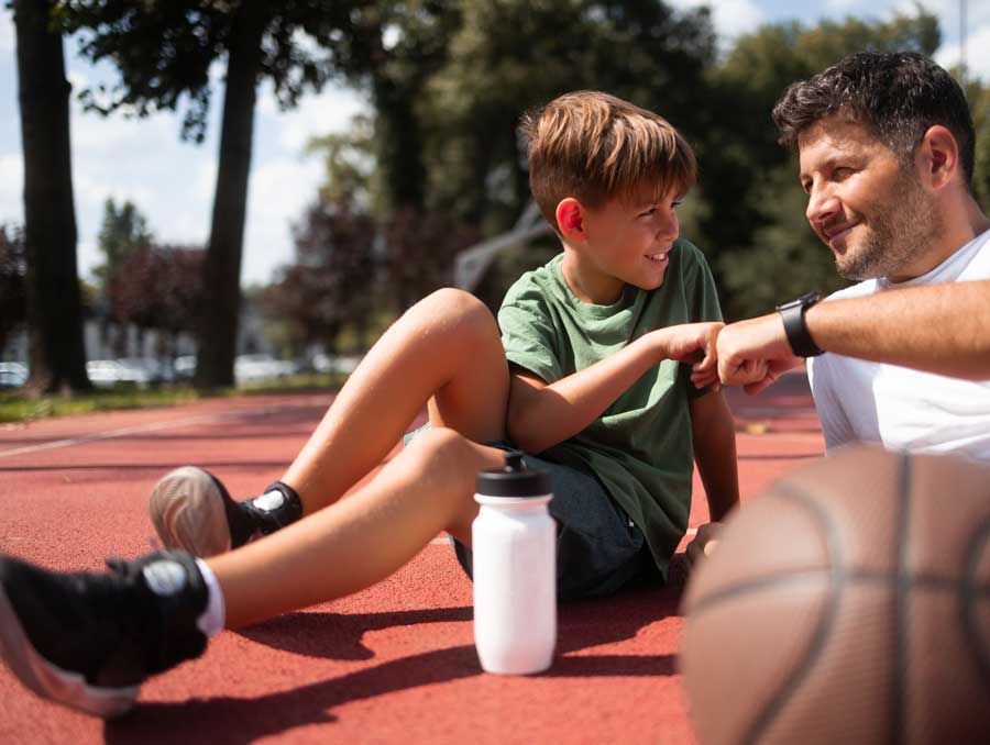 A father and son bump fists after a basketball game. They're sprawled on the court next to a water bottle.