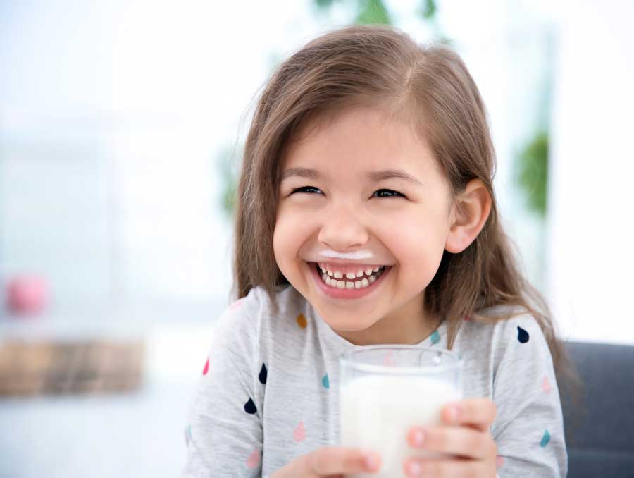 A girl giggles while holding a glass of milk. Her smile is adorned by a milk moustache.