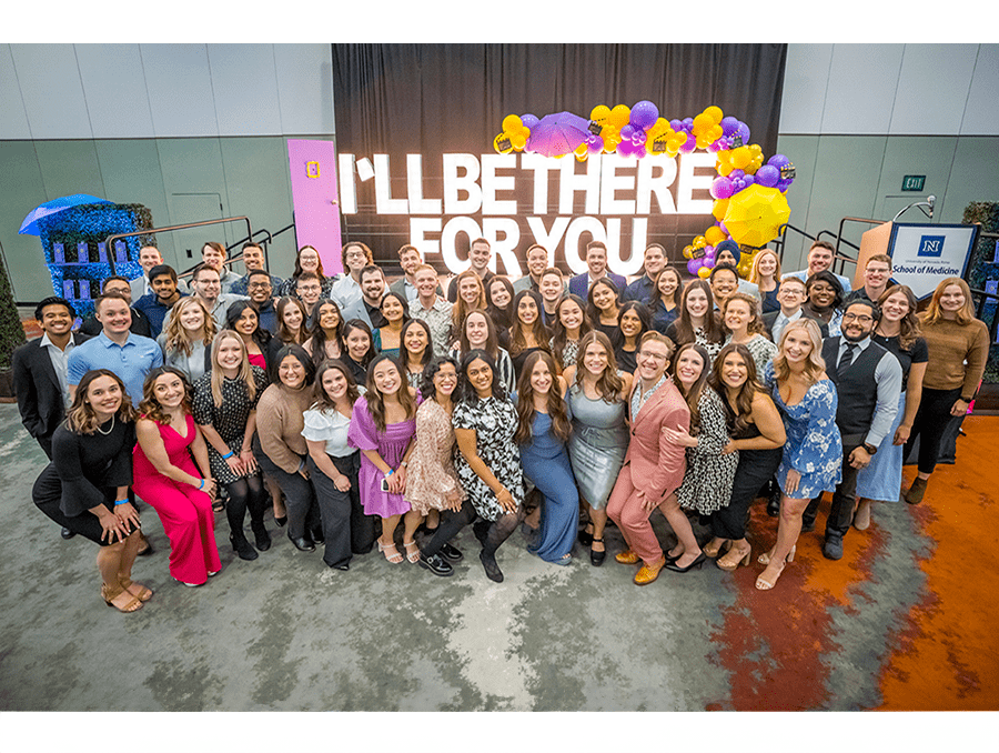 Medical students pose in front of the stage for a silly group photo, with "I'll Be There For You" sign in the background.