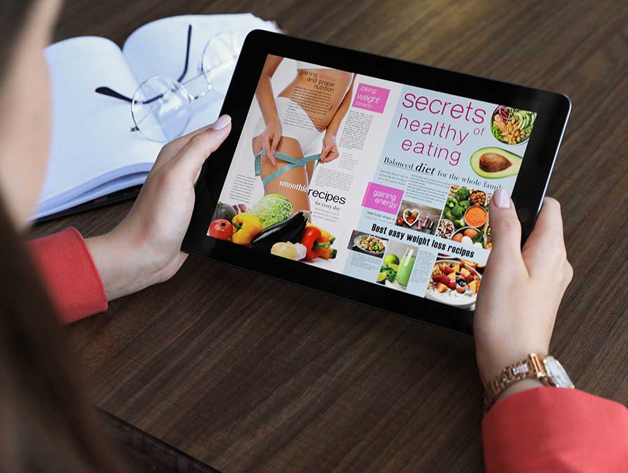 A woman holding an iPad displaying a website on dieting. Nearby are her reading glasses and a notebook.