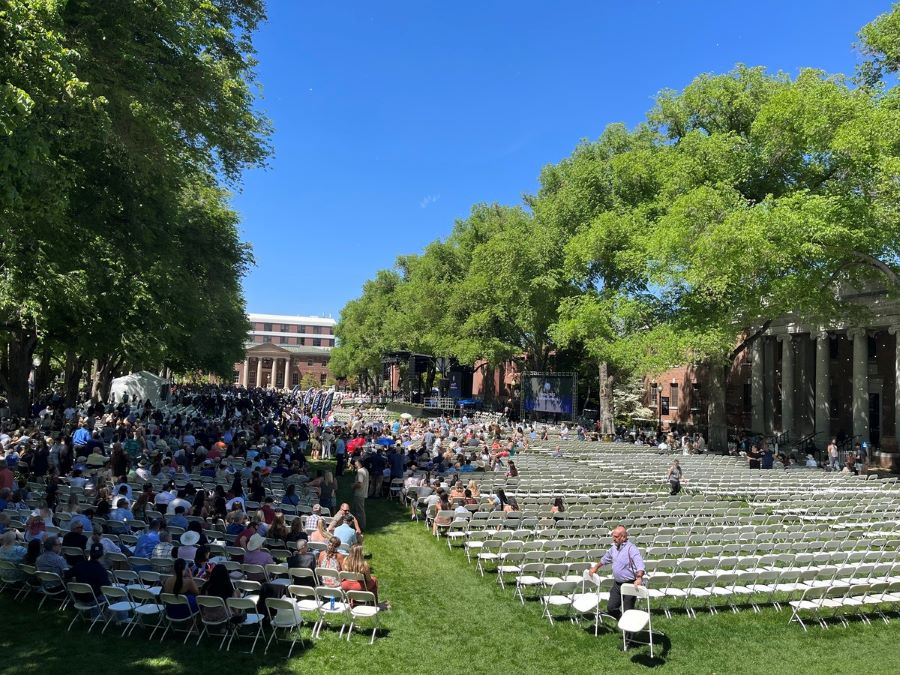 A crowd gathers on the quad to prepare for commencement ceremonies. Rows of empty chairs are set up.
