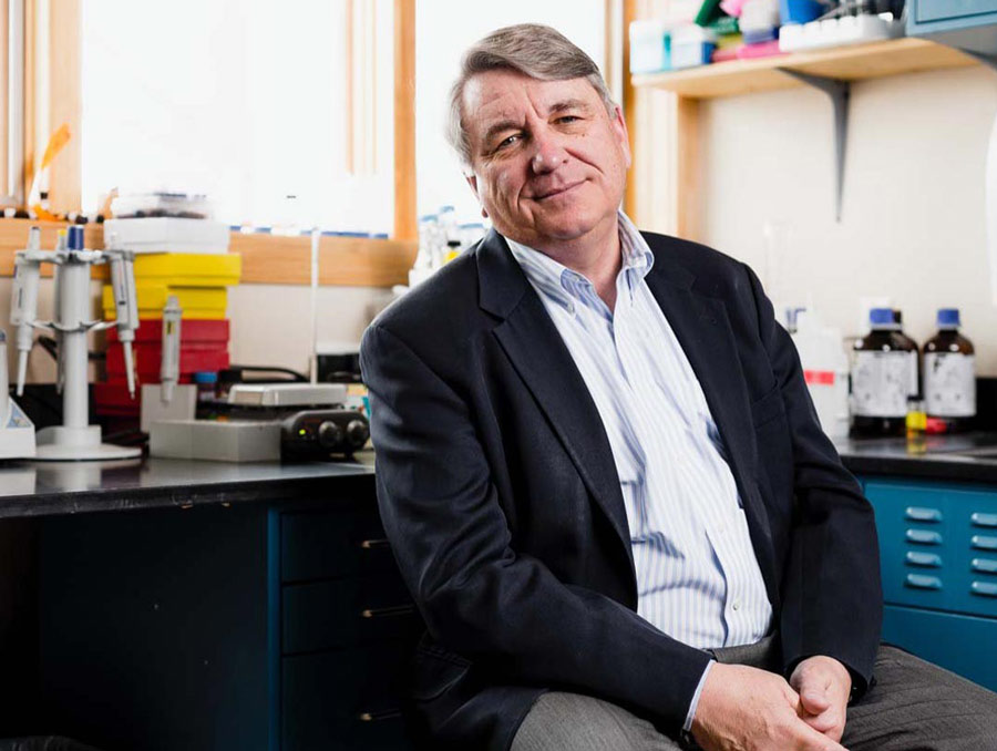 Paul Alan Cox sits in a lab space with bottles of chemicals and pipettes behind him.