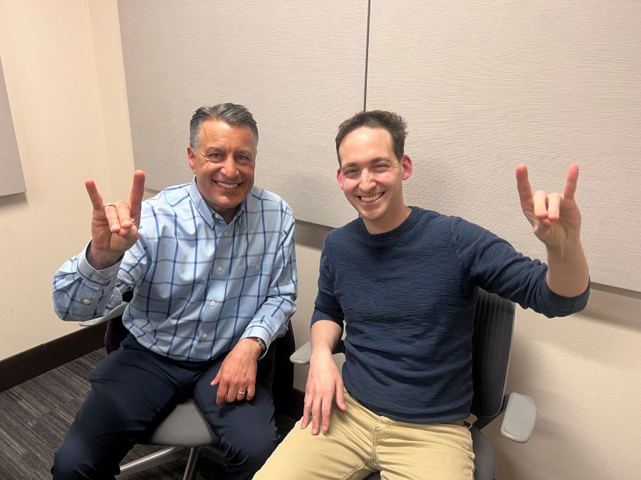 Brian Sandoval sitting next to Thomas White in the podcasting studio holding up Wolf Pack hand signs.
