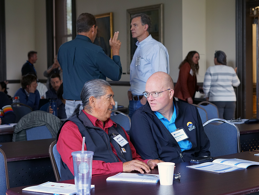 Two men sitting at a table talking with people behind them talking in groups.