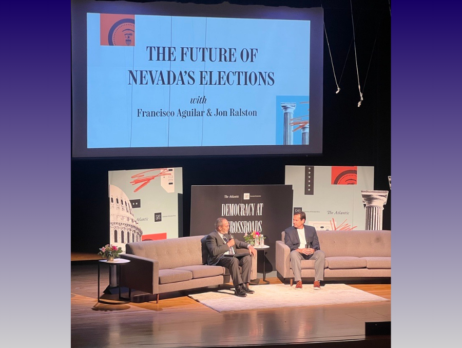 John Ralston sits next to Francisco Aguliar on stage at the Democracy at a Crossroads event with couches on stage and signage about the Atlantic and the event behind them.