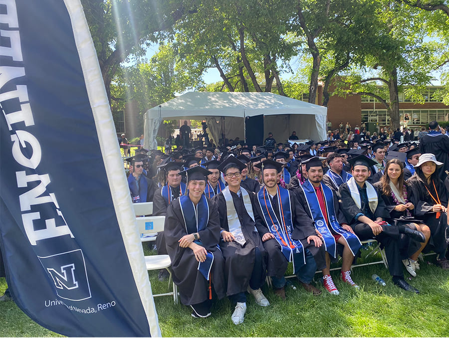 Engineering students in graduation robes and caps sitting on lawn chairs on the quad next to a banner that reads "Engineering"