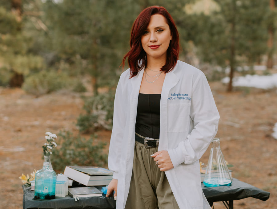 Hailey wearing her lab coat standing in front of a table in the woods with lab supplies, books, and flowers.