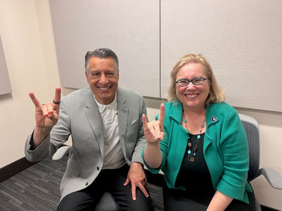 Brian Sandoval sitting next to Muge Akpinar in the podcasting studio holding up Wolf Pack hand signs.