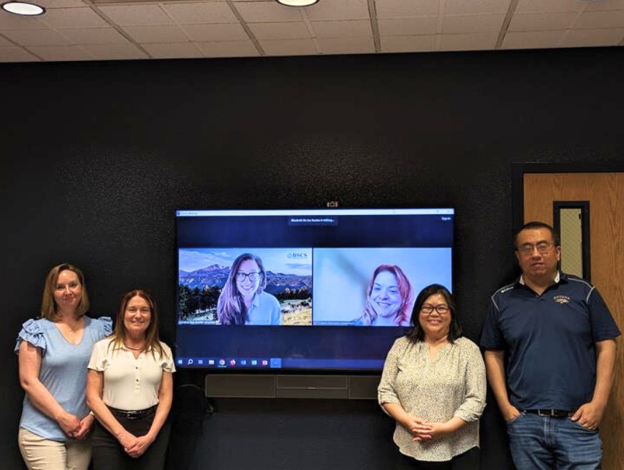 Four faculty standing next to a monitor with two people on the screen.