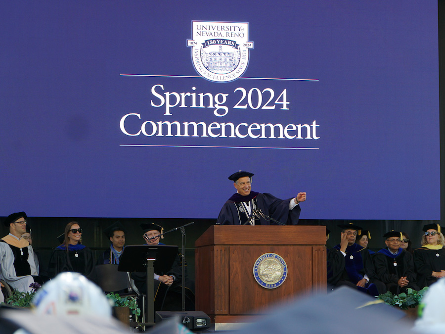 President Sandoval stands at the podium in graduation regalia while giving his speech during the Spring 2024 Commencement Ceremonies. The sesquicentennial logo appears behind him on a PowerPoint along with "Spring 2024 Commencement."