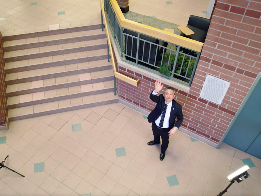 Dean Yun waving at a drone that is flying up above him in the middle of the Reynolds School atrium.