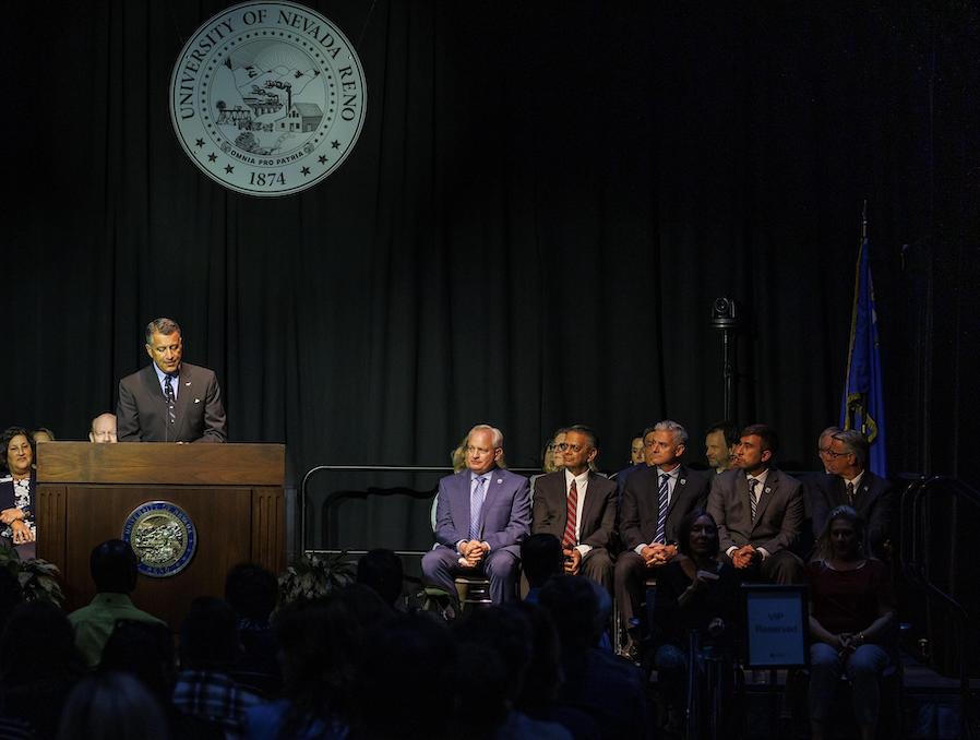 President Sandoval stands on stage with a group of University leadership sitting behind him while he speaks from a podium. The Seal of the University of Nevada, Reno is behind him.