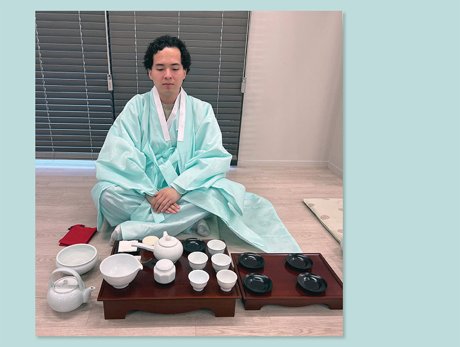 A man wearing a light green robe kneels in front of a tea setting on a wooden tray.