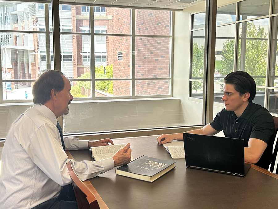 Two individuals sitting at a table in a library, discussing over books and a laptop, with large windows overlooking buildings in the background.