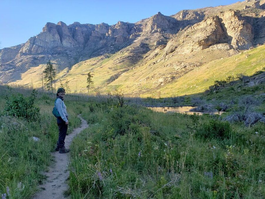 A smiling woman stands on a dirt path lined with green bushes, with mountains in the background.