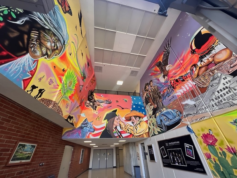 Three walls in Cain Hall depicting various scenes including an elderly woman with butterflies and a rose with green light emanating from it, a woman with a graduation cap being adjusted by an older ranch man, a wolf, the American flag, desert cacti, an eagle, a classic Volkswagon bug, an astronaut with stars, and a church.
