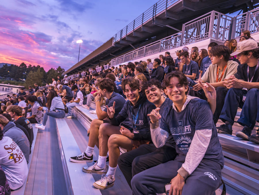 Mackay stadium filled with students during sunset - many students wear Nevada t-shirts and some smile and hold up the Wolf Pack hand sign.