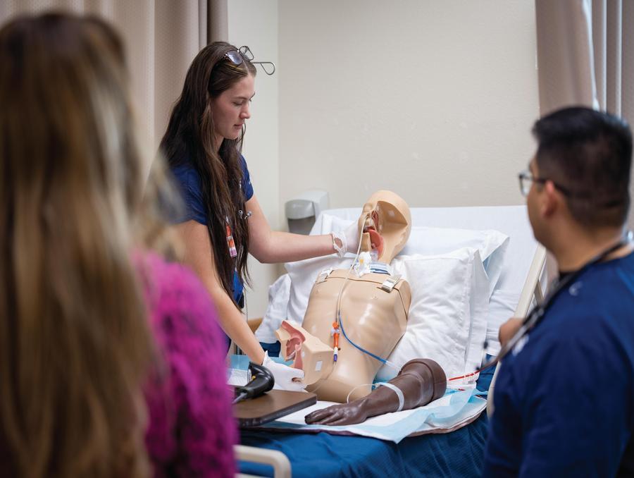 A nursing student practices on a simulated hospital bed with a dummy patient lying in the bed while more students look on, observing.