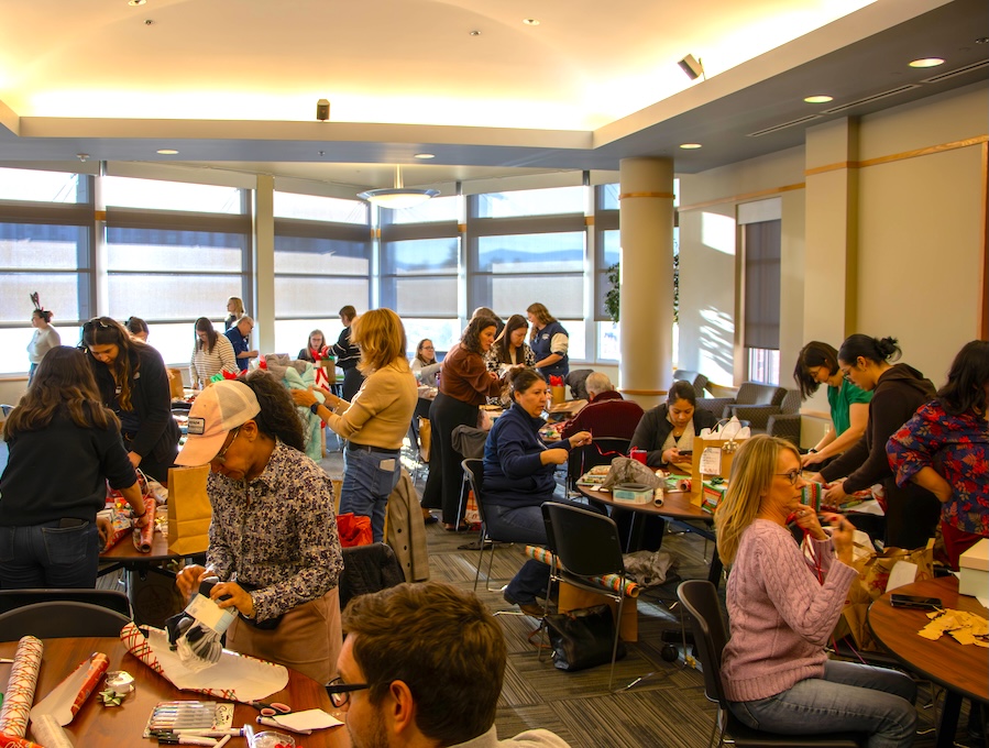 A large group of employees all wrapping presents as they talk together at tables.