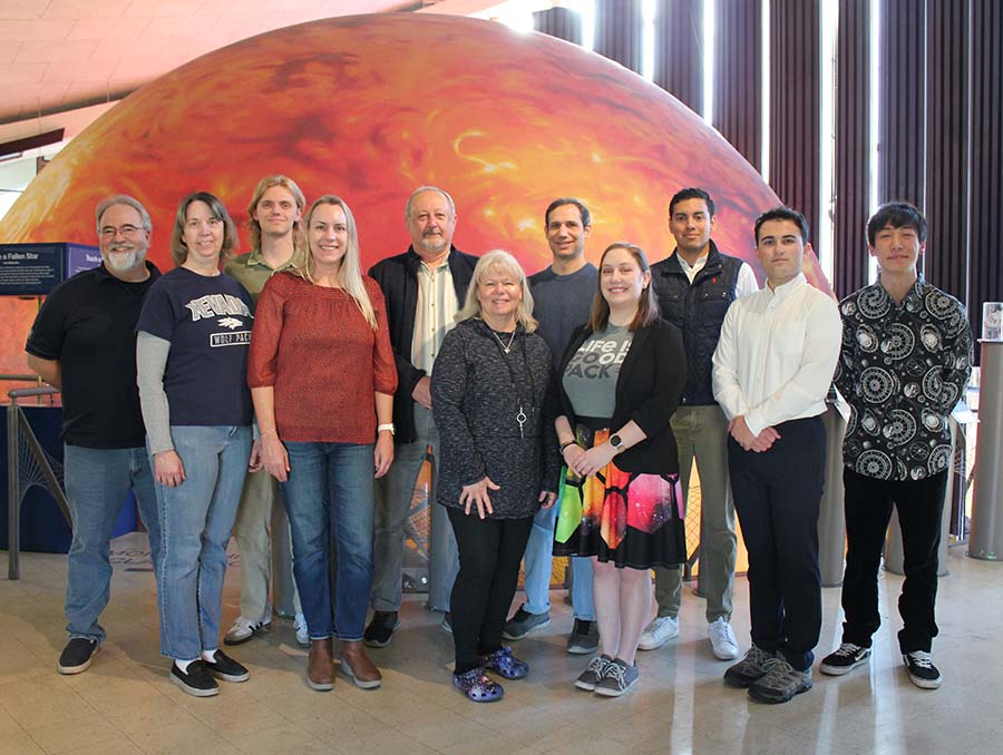 Eleven people pose inside a planetarium, in front of a large model of a sun.