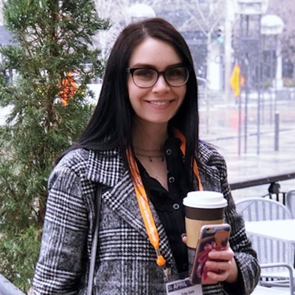 A woman wearing a coat and holding a paper coffee cup stands in front of a tree, with a street in the background.
