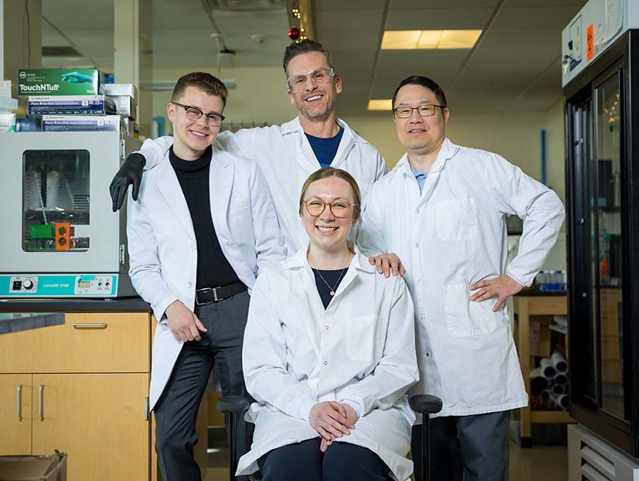 Samuel Lee, Travis Stewart, Hannah Schultz, and Riley Gillis in a laboratory wearing lab coats and smiling at the camera.