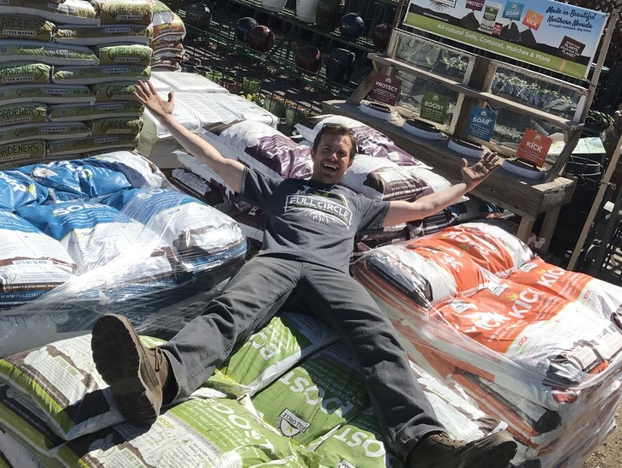 A smiling man wearing a Full Circle Compost shirt lies playfully on top of stacked compost bags outdoors, surrounded by gardening soil products and display shelves.