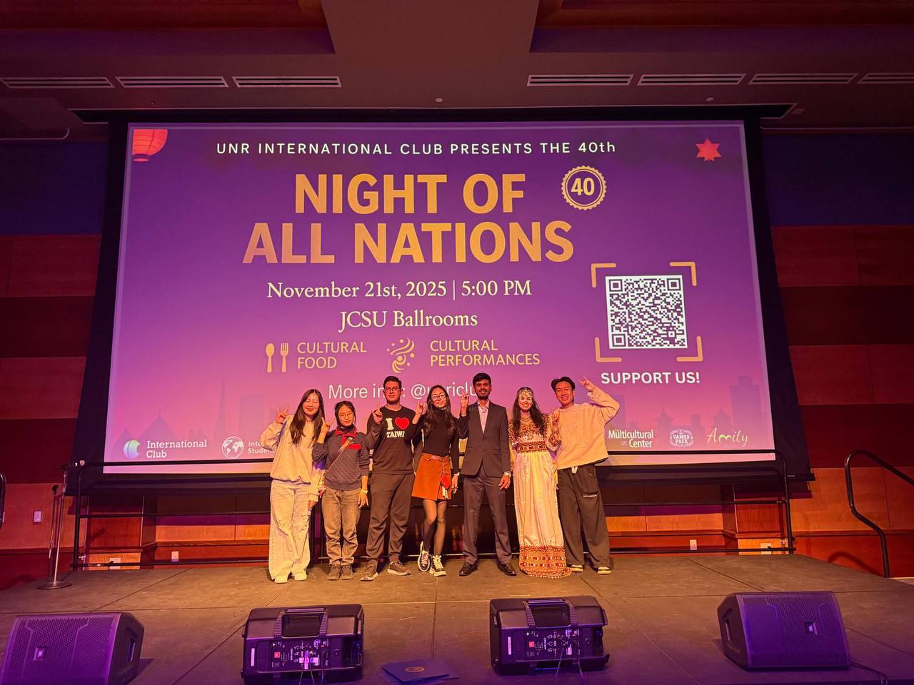 A group of 7 students, some wearing cultural clothing, stand on stage in front of the Night of All Nations PowerPoint, holding up Wolf Pack hand signs..