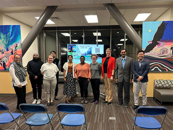 A group of 11 people, students, faculty and presenters, stand together in a campus event space with colorful artwork on the walls and a large presentation screen in the background.