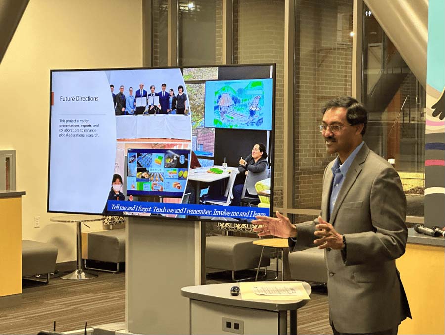 A man in a gray suit stands and gestures while speaking in front of a large screen displaying slides with photos and the heading “Future Directions.” The images on the slide include group photos and student work.