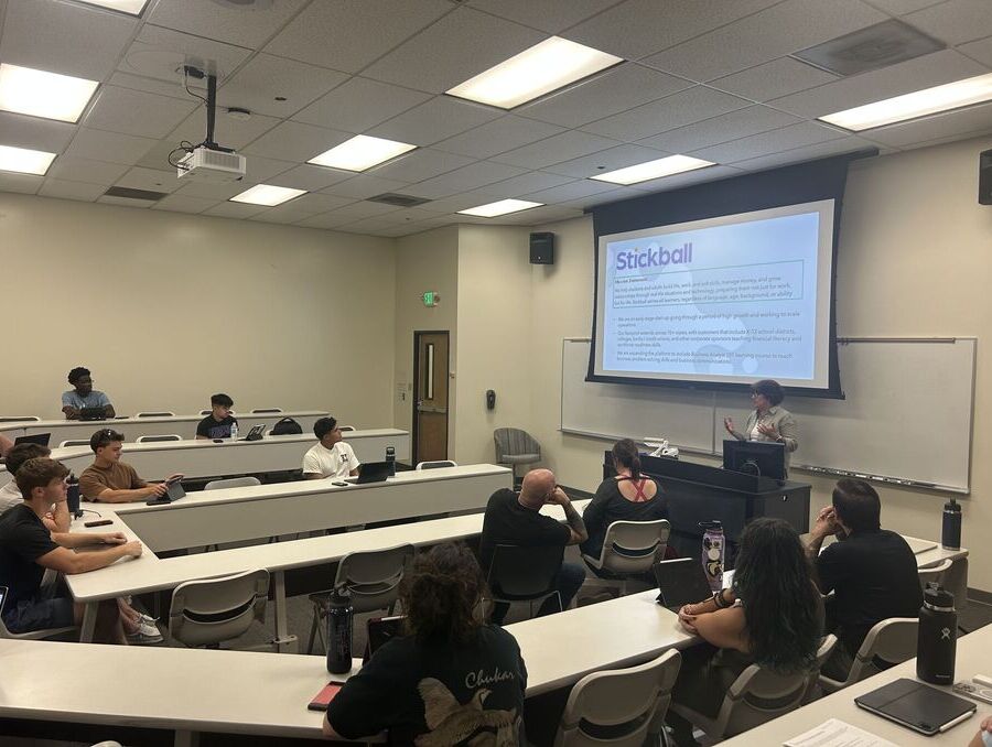 Students in the MGT 321 service-learning class sit at desks in a classroom while a community partner presents a slide about Stickball on a projector screen at the front of the room.